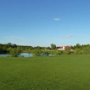 Golfplatz Achim Grüne Landschaft mit Golfplatz im Vordergrund, einem kleinen See und einem roten Gebäude im Hintergrund.