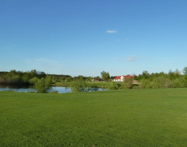 Golfplatz Achim Grüne Landschaft mit Golfplatz im Vordergrund, einem kleinen See und einem roten Gebäude im Hintergrund.
