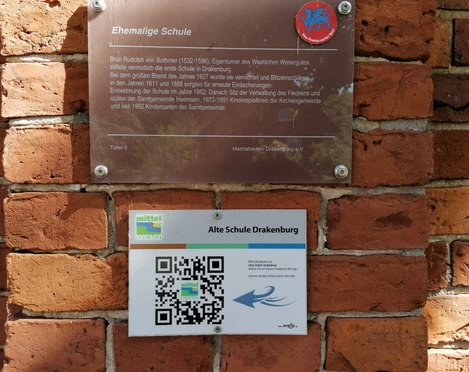 Alte Schule Drakenburg Eine Infotafel aus Metall und ein Schild mit QR-Code sind an einer Backsteinmauer angebracht.