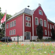 Amtshof 1 Bruchhausen-Vilsen Amtshof 1 in Bruchhausen-Vilsen, ein rotes Gebäude mit weißen Fensterrahmen und Blumenwiese davor.
