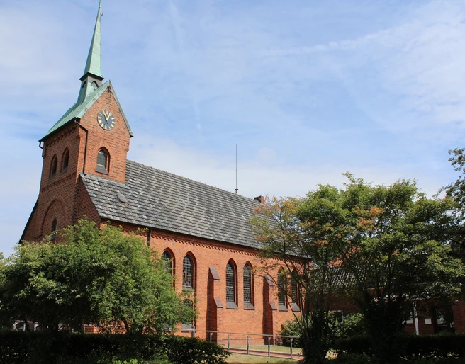 Kirche Bruchhausen Backsteinkirche mit Spitzdach und Turm im Dorf Bruchhausen; von Bäumen umgeben, blauer Himmel.