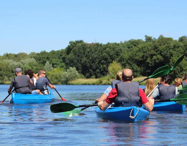 Kanutour Mittelweser Eine Gruppe von Menschen in blauen Kanus paddelt über einen ruhigen Fluss, umgeben von grünen Bäumen.