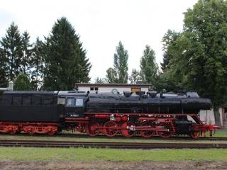 Museumslok Weyhe Eine historische Dampflokomotive steht auf Schienen, umgeben von Bäumen und einem Gebäude im Hintergrund.