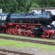 Historische Dampflok im Freien, schwarz mit roten Rädern, umgeben von Bäumen und einer Hütte.