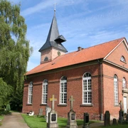 Kirche Oiste Backsteinkirche mit Spitzdach und Glockenturm, umgeben von Bäumen und Grabsteinen unter blauem Himmel.