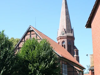 Ein Kirchturm mit Uhr ragt hinter grünen Bäumen und einem roten Backsteingebäude empor.