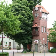 Feuerwehrturm aus rotem Backstein mit weißen Fenstern und markantem Balkon, umgeben von Bäumen.