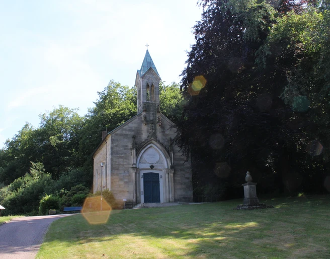 Eine historische Kapelle mit gotischen Elementen, umgeben von üppigem Grün und einem Gedenkstein.
