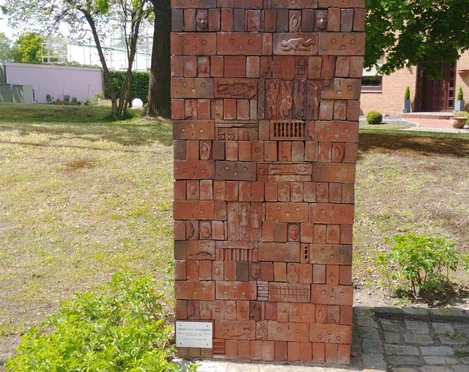 Gegen das Vergessen Ein hohes, rechteckiges Mosaikdenkmal aus roten Ziegeln mit eingravierten Symbolen und Figuren.