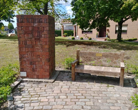 Gegen das Vergessen Gedenksäule aus roten Ziegeln neben einer hölzernen Bank auf gepflastertem Platz, umgeben von Bäumen.
