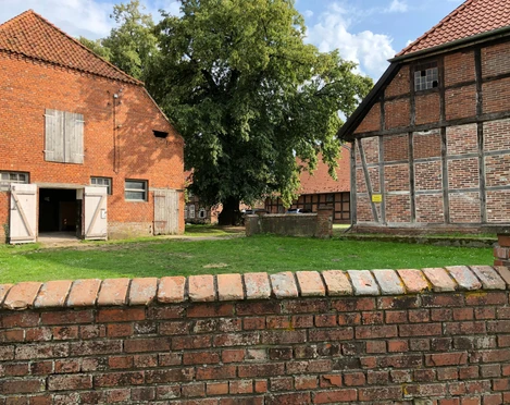Gut Oenigstedt Roter Backsteinbau und Fachwerkgebäude rahmen einen grünen Hof mit großen schattenspendenden Bäumen.