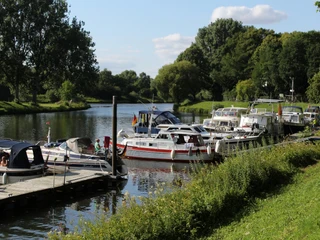 Hafen Nienburg Am Flussufer liegen Boote an einem Holzsteg vertäut, umgeben von üppigem Grün und Bäumen.