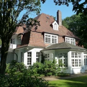Haus Hünenburg Ein elegantes historisches Gebäude mit großem Dach und hellen Fenstern, umgeben von üppigem Grün.