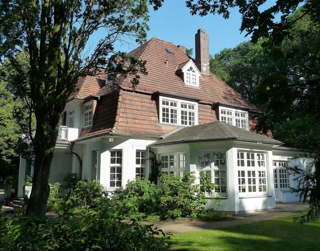 Haus Hünenburg Ein elegantes historisches Gebäude mit großem Dach und hellen Fenstern, umgeben von üppigem Grün.