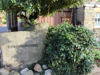 Stein mit Inschrift "Heimatmuseum" vor Fachwerkgebäude umgeben von Sträuchern und Gehweg.