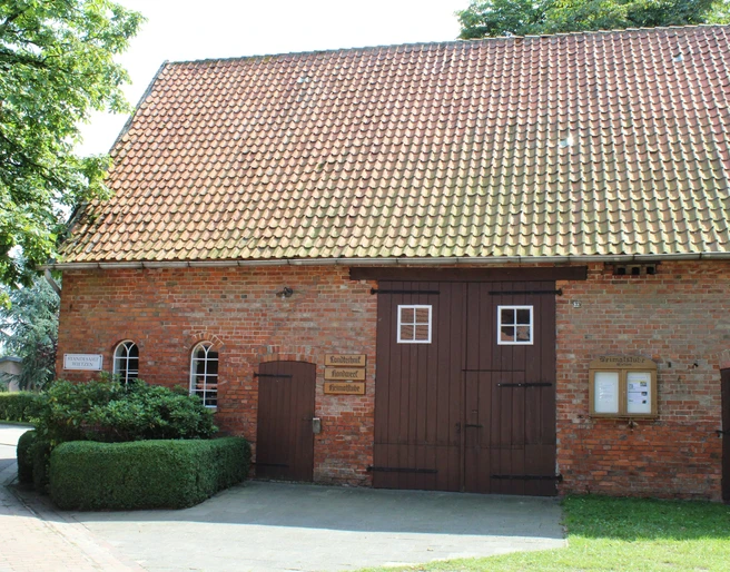 Heimatstube Wietzen Ziegelsteinbau mit Satteldach in Wietzen, Eingang mit Doppeltür, umgeben von grüner Hecke.