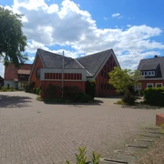 Kath. Kirchengemeinde Heilige Familie Rote Backsteinkirche im Licht der Sonne, umgeben von Bäumen, mit blauem Himmel und weißen Wolken.