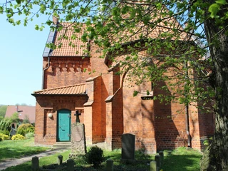 Backsteinkirche mit grünem Tor, umgeben von Bäumen und Gräbern, im ländlichen Magelsen.