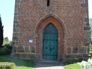Backsteinkirche in Magelsen mit grünem Holzportal und gotischem Spitzbogen, umgeben von gepflegtem Grün.