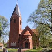 Backsteinkapelle mit spitzem Turm und Uhr, umgeben von Bäumen vor strahlend blauem Himmel.