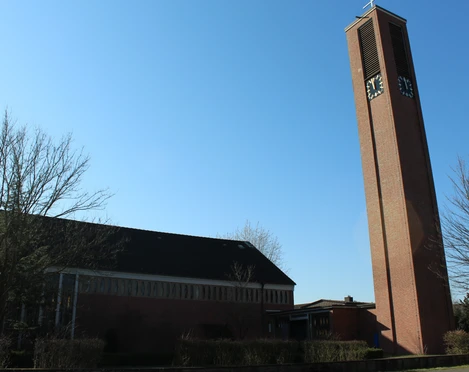 Backsteinkirche mit hohem Glockenturm, Uhr an der Spitze, blauer Himmel im Hintergrund, Bäume ohne Blätter.