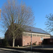 Backsteinkirche mit markantem Turm und Uhr, umgeben von kahlen Bäumen, an einem klaren, blauen Himmel.