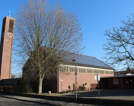 Backsteinkirche mit markantem Turm und Uhr, umgeben von kahlen Bäumen, an einem klaren, blauen Himmel.