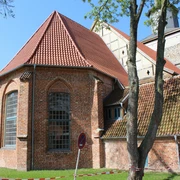 Backsteinkirche mit roten Dachziegeln und gotischen Spitzbogenfenstern, umgeben von Bäumen und Wiese.