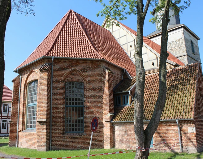 Backsteinkirche mit roten Dachziegeln und gotischen Spitzbogenfenstern, umgeben von Bäumen und Wiese.