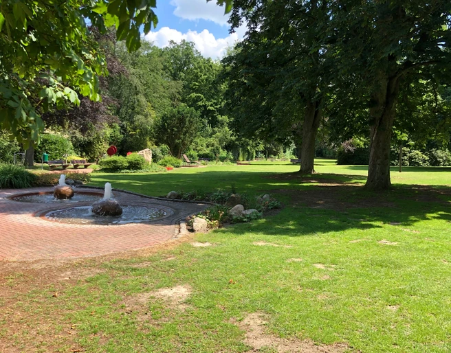 Kurpark Ein gepflegter Kurpark mit sprudelndem Brunnen, üppigen Bäumen und weitläufigen Rasenflächen.