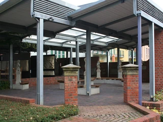 Eingang Ein überdachter Eingangsbereich mit roten Backsteinsäulen, Skulpturen und gläsernen Dachpaneelen.