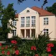 Kreismusikschule - Lindemann'sches Schloß Quadratisch-vornehmes Schlossgebäude mit roten Dachziegeln, umgeben von grünen Bäumen und roten Blumen.