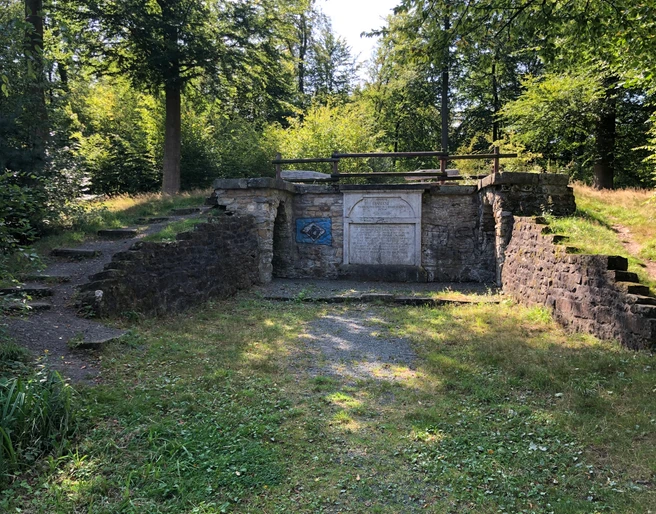 Ruinen einer historischen Steinmauer inmitten eines lichten Waldes an einem sonnigen Tag.