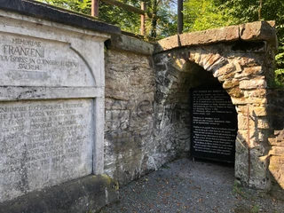 Historische Steinmauer mit lateinischer Inschrift und einem dunklen Eingang, umgeben von grünem Laub.