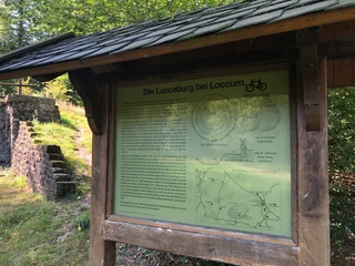 Informationsstand im Grünen mit Text zur Luccaburg bei Loccum, daneben alte Steinstrukturen.