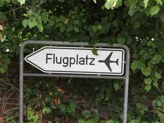 Wegweiser zum Flugplatz mit Abbildung eines Flugzeugs vor einer dichten Laubhecke.