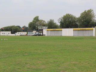Grüne Wiese mit Hangars und Kontrollturm eines kleinen Flugplatzes unter bewölktem Himmel.