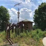 Marschbodenglück – Schibboleth Skulptur eines Widders aus Stahl, umgeben von Wildblumen und einem hölzernen Pavillon im Marschland.