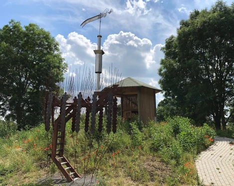 Marschbodenglück – Schibboleth Skulptur eines Widders aus Stahl, umgeben von Wildblumen und einem hölzernen Pavillon im Marschland.
