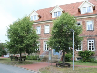 Mehrgenerationenhaus Ein Ziegelbau mit rotem Dach, Treppenzugang und Sitzbänken, umgeben von Bäumen und Strauchwerk.