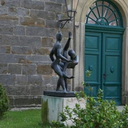 Skulptur "Paartanz" Zwei tanzende Figuren aus Bronze vor einem historischen Gebäude mit grünem Holztor und Pflanzen.