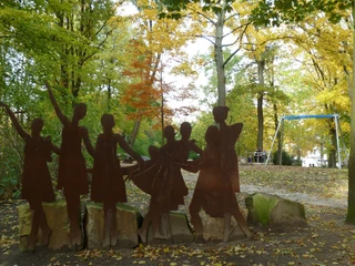 Rathauspark Achim Eine rostfarbene Stahlskulptur von tanzenden Menschen in einem herbstlichen Park mit bunten Blättern.