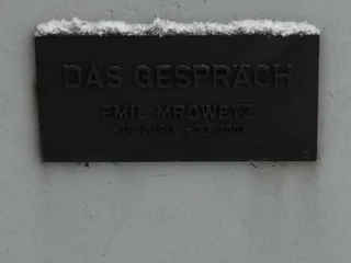 Schild an der Skulptur "Das Gespräch" Tafel mit der Inschrift "Das Gespräch" von Emil Mrowetz, beleuchtet durch Schneeakzente oben.
