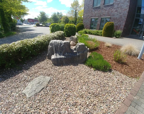 Die Liegende Steinskulptur einer liegenden Figur umgeben von Kiesbeet und grüner Bepflanzung vor einem Gebäude.