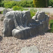 Die Liegende Steinskulptur mit liegenden Figuren und Gesicht auf einem Kiesbett, umgeben von Pflanzen und Gehweg.