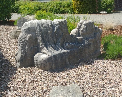 Die Liegende Steinskulptur mit liegenden Figuren und Gesicht auf einem Kiesbett, umgeben von Pflanzen und Gehweg.