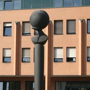 Flora II Skulptur mit kugelförmiger Spitze vor einem Backsteingebäude mit zahlreichen quadratischen Fenstern.