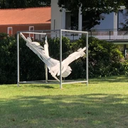 Gefallen Eine skulpturale Darstellung von verknoteten weißen Stoffstreifen in einem quadratischen Metallrahmen auf einer grünen Rasenfläche. Im Hintergrund sind dichtes Gebüsch und ein modernes Gebäude mit rotem Ziegeldach und Balkon zu sehen.