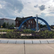 Skulptur Kreisel Hoya Skulptur eines blauen Rades auf einem Kreisverkehr in Hoya, umgeben von Blumen und Industriegebäuden.