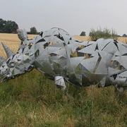Skulptur Nashorn Nashornskulptur aus Metallfragmenten vor einem Getreidefeld und Bäumen, eingehüllt von wilder Vegetation.